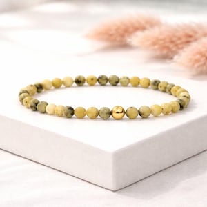 Könnte beinhalten: Ein Perlenarmband bestehend aus kleinen, runden, gesprenkelten gelben und grünen Steinperlen, die in einem Kreis angeordnet sind, akzentuiert durch eine einzelne, glatte, polierte goldfarbene Metallperle nahe der Mitte.