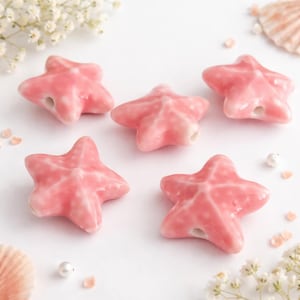 Könnte beinhalten: Fünf rosa Keramik-Seestern-Perlen mit glänzendem Finish. Jeder Seestern hat ein Loch zum Auffädeln. Die Perlen sind auf einer weißen Oberfläche mit dekorativen Elementen angeordnet.