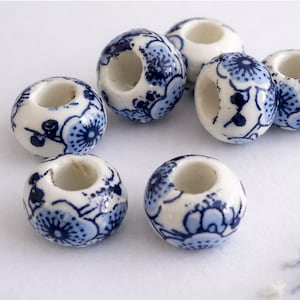 Könnte beinhalten: Eine Ansammlung weißer Keramikperlen mit blauem Blumenmuster. Jede Perle hat ein zylindrisches Loch in der Mitte. Die Perlen sind auf einer weißen Oberfläche angeordnet, um ihr aufwendiges Design zu zeigen.