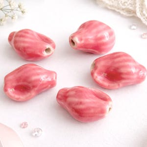 Könnte beinhalten: Fünf rosa Keramikperlen mit glänzendem Finish. Jede Perle hat eine einzigartige, organische Form mit einem Loch zum Auffädeln. Die Perlen haben ein gestreiftes Muster in einem helleren Rosaton, wodurch ein strukturiertes Aussehen entsteht. Ideal für die Schmuckherstellung.