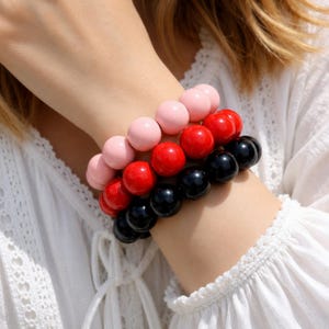 Könnte beinhalten: Ein Set aus drei gestapelten Perlenarmbändern mit glatten, glänzenden, kugelförmigen Perlen. Die Armbänder sind in verschiedenen Farbschichten angeordnet und bestehen aus einer Reihe hellrosa Perlen, einer Reihe leuchtend roter Perlen und einer Reihe schwarzer Perlen.