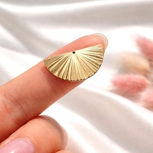 Könnte beinhalten: Ein goldfarbenes, fächerförmiges Schmuckteil mit strukturierter, geriffelter Oberfläche. Das halbkreisförmige Design hat oben ein kleines Loch zur Befestigung. Das Schmuckteil wird zwischen zwei Fingern vor einem weichen, weißen Hintergrund gehalten.