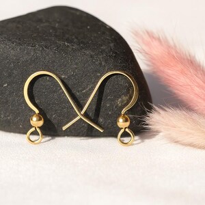 May include: A pair of gold-colored earring hooks. Each hook features a small, round bead and a looped bottom. The hooks are displayed against a dark gray stone and a backdrop of pink and white decorative elements.