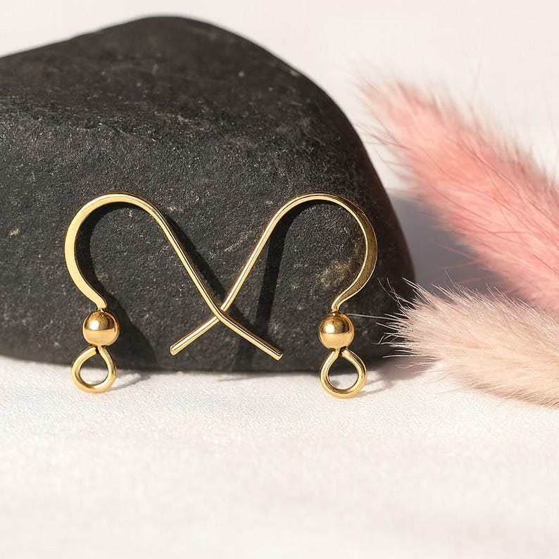 May include: A pair of gold-colored earring hooks. Each hook features a small, round bead and a looped bottom. The hooks are displayed against a dark gray stone and a backdrop of pink and white decorative elements.