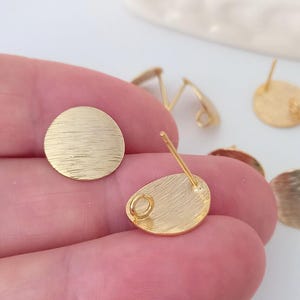 Könnte beinhalten: Goldfarbene Ohrringrohlinge mit gebürsteter Oberfläche. Die Rohlinge sind rund und haben einen Stift mit einer Öse auf der Rückseite.