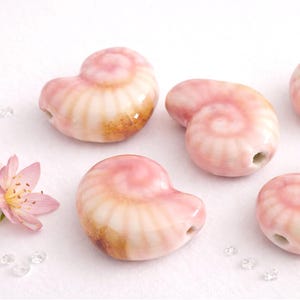 Könnte beinhalten: Mehrere keramische Muschelkernperlen in Rosa- und Cremetönen sind auf einer weißen Oberfläche angeordnet. Die Perlen haben ein Spiraldesign und ein Loch zum Auffädeln. Eine rosa Blume und kleine, klare Kristalle sind ebenfalls vorhanden.