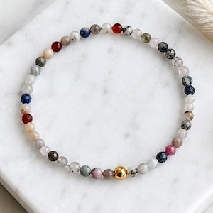 Könnte beinhalten: Ein zartes Perlenarmband mit kleinen, facettierten, mehrfarbigen Edelsteinperlen in Rot-, Blau-, Grau-, Weiß- und Rosatönen. Der kreisförmige Strang wird durch eine einzelne, glatte, polierte goldfarbene Metallperle im unteren Bereich akzentuiert.