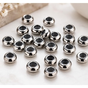 Könnte beinhalten: Eine Ansammlung kleiner, runder, metallisch silberner Perlen mit einer glatten, polierten Oberfläche. Jede Perle hat ein zentrales kreisförmiges Loch, was eine gleichmäßige, donutartige Form erzeugt. Die Perlen liegen verstreut auf einer hellen Fläche.