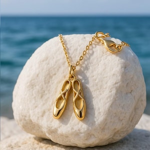 Könnte beinhalten: Eine goldfarbene Halskette mit Anhänger, die ein Paar Ballettschuhe zeigt. Der Anhänger hängt an einer filigranen Kette mit einem Verschluss. Die Halskette wird auf einem weißen Felsen mit verschwommenem Ozean im Hintergrund präsentiert.