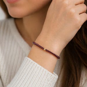 Könnte beinhalten: Ein Armband mit kleinen, dunkelroten Perlen und einer einzelnen goldfarbenen Perle. Das Armband wird am Handgelenk getragen und unterstreicht seine schlichte Eleganz. Die Perlen haben einen Durchmesser von etwa 0,5 cm.