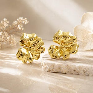 Könnte beinhalten: Ein Paar goldfarbene, blattförmige Ohrringe. Die Ohrringe haben eine detaillierte, strukturierte Oberfläche und ein glänzendes Finish. Sie werden auf einer hellen, strukturierten Oberfläche mit einem weichen, neutralen Hintergrund präsentiert.