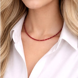 Könnte beinhalten: Eine zarte, rote Perlenkette. Die Kette besteht aus kleinen, runden Perlen, die einen durchgehenden Strang bilden. Die Kette wird über einem weißen Hemd getragen.
