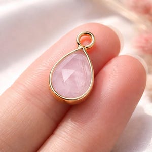 Könnte beinhalten: Ein tropfenförmiger Anhänger mit einem facettierten, hellrosa Edelstein, der in eine goldfarbene Fassung gefasst ist. Der Anhänger hat oben eine kleine Schlaufe zum Anbringen an einer Kette. Der Hintergrund ist ein weiches, verschwommenes Rosa.