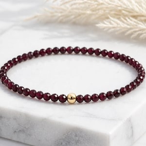 Könnte beinhalten: Ein zartes Perlenarmband mit kleinen, facettierten, tiefroten Granat-Edelsteinen in einem durchgehenden kreisförmigen Strang, akzentuiert durch eine einzelne, glatte, polierte goldfarbene Kugelperle in der Mitte des Designs.
