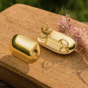 Könnte beinhalten: Zwei goldfarbene Schmuckstücke. Eines ist ein solider, abgerundeter Rechteck mit einem kleinen Loch. Das andere ist eine ovale Schale mit einem Pfosten und einem Ring. Die Artikel befinden sich auf einer Holzoberfläche.