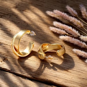 Könnte beinhalten: Ein Paar goldfarbene Creolen mit einem verdrehten Design. Die Ohrringe liegen auf einer Holzoberfläche, im Hintergrund getrocknete Blumen. Die Ohrringe haben einen Stecker und einen Butterfly-Verschluss.