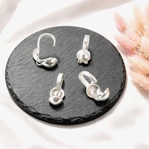 Könnte beinhalten: Vier silberfarbene Schmuckelemente aus Metall, speziell Quetschkalotten oder Perlenkappen, angeordnet auf einer runden schwarzen Schieferplatte. Jedes Teil verfügt über eine kleine Öse an der Oberseite und eine hohle, abgerundete Basis.