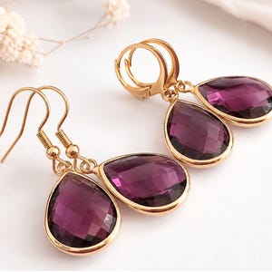 Könnte beinhalten: Ein Paar goldfarbene Ohrringe mit facettierten, birnenförmigen violetten Kristallen. Ein Ohrring hat einen Hakenverschluss, während der andere mit einem Klappbügel versehen ist. Die tiefvioletten Steine sind in polierte Goldfassungen gefasst.