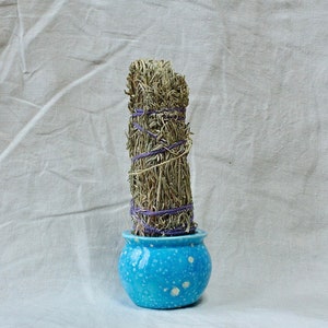 May include: A cylindrical bundle of dried, light brown herbs tightly wrapped with purple string, standing upright inside a small, round, bright blue ceramic pot with a speckled white finish and a smooth, glossy surface texture.