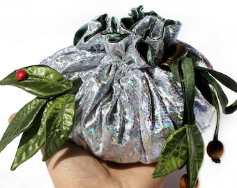 May include: A silver iridescent drawstring bag with green fabric leaves and beads. The bag is held in a hand and there are two dice on a white surface.