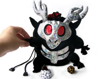 May include: Black plush dice bag shaped like a skull with silver and iridescent horns. The bag has red eyes and a drawstring closure. Two dice are in front of the bag, one white and one black.