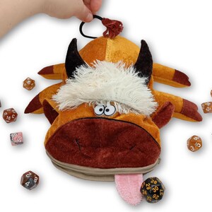 May include: A plush, orange and brown cow-shaped dice bag with black horns, white fur, and a pink tongue. The bag has a zipper closure and a black loop. Several multi-sided dice in various colors surround the bag.