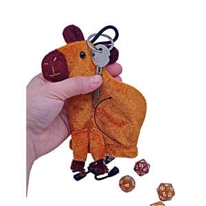 May include: A plush brown and red camel keychain with a metal key attached. The camel is holding a small black pouch with a drawstring. There are four brown dice with white numbers on them scattered around the camel.