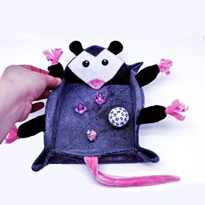 May include: A plush grey and black possum-shaped dice rolling tray with pink accents. The tray is open and has dice inside, including a white d20.