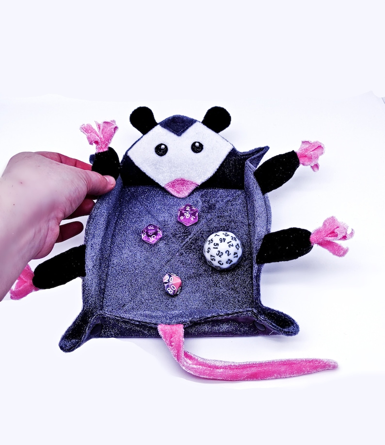 May include: A plush grey and black possum-shaped dice rolling tray with pink accents. The tray is open and has dice inside, including a white d20.