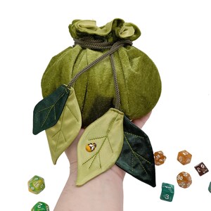 Puede incluir: Bolsa de dados de terciopelo verde con cierre de cord&oacute;n y dos hojas de sat&eacute;n verde con un peque&ntilde;o adorno de mariquita. La bolsa est&aacute; siendo sostenida por una mano y hay varios dados esparcidos sobre una superficie blanca.