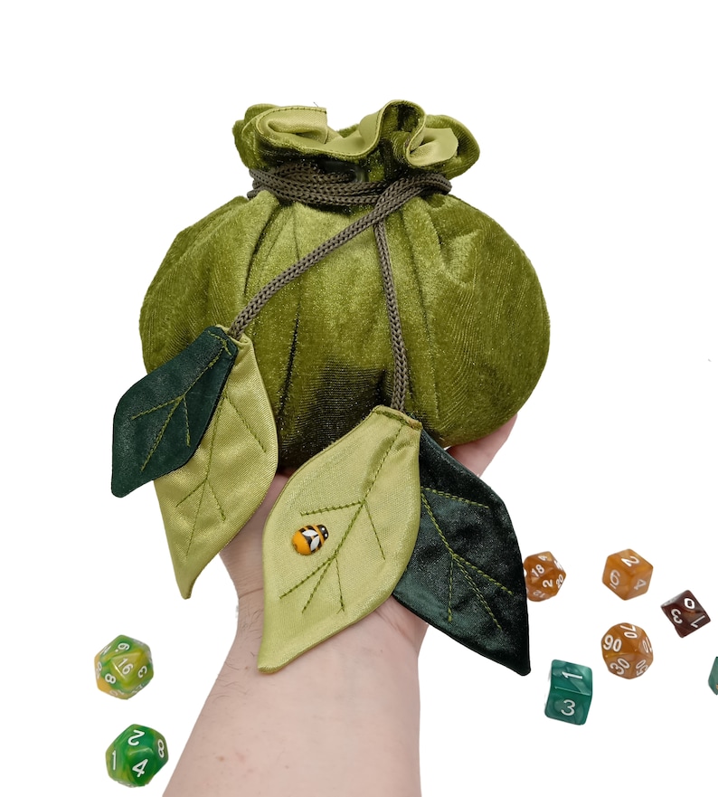 Puede incluir: Bolsa de dados de terciopelo verde con cierre de cord&oacute;n y dos hojas de sat&eacute;n verde con un peque&ntilde;o adorno de mariquita. La bolsa est&aacute; siendo sostenida por una mano y hay varios dados esparcidos sobre una superficie blanca.