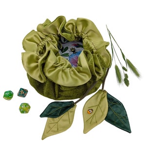 Puede incluir: Bolsa de dados de terciopelo verde con cierre de cord&oacute;n y dos hojas de tela verde con dise&ntilde;o de abeja. La bolsa est&aacute; forrada con un tejido de sat&eacute;n verde claro y tiene una peque&ntilde;a tarjeta con estampado floral en su interior. Hay tres dados verdes sobre un fondo blanco.
