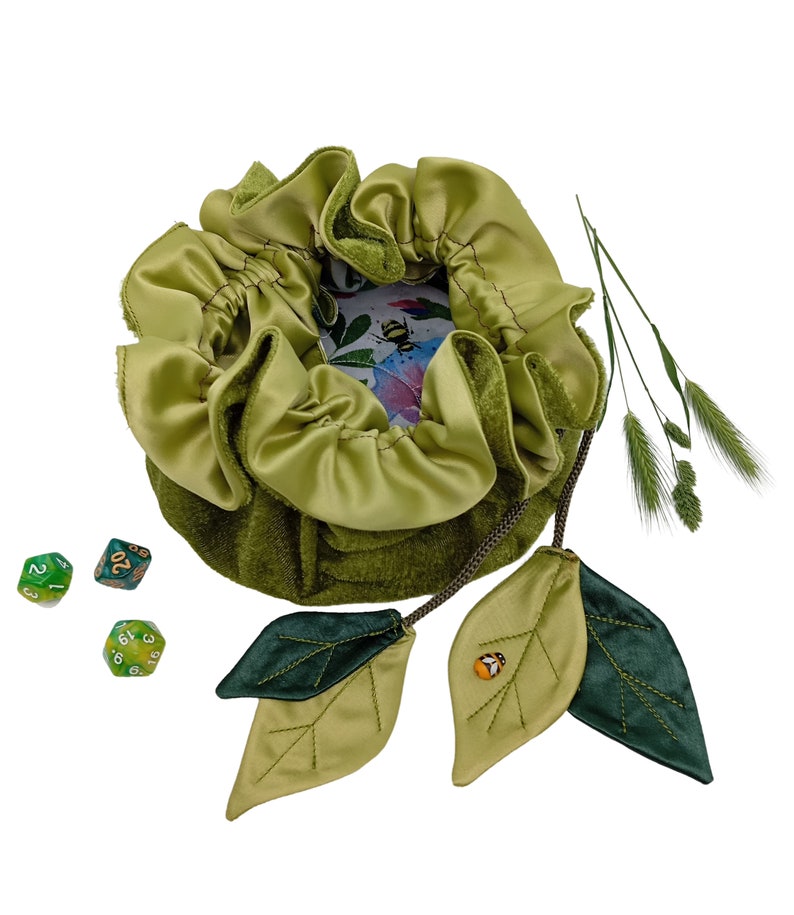 Puede incluir: Bolsa de dados de terciopelo verde con cierre de cord&oacute;n y dos hojas de tela verde con dise&ntilde;o de abeja. La bolsa est&aacute; forrada con un tejido de sat&eacute;n verde claro y tiene una peque&ntilde;a tarjeta con estampado floral en su interior. Hay tres dados verdes sobre un fondo blanco.