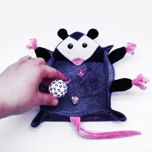 May include: A plush grey and pink opossum dice tray with a pink nose and black eyes. The tray is filled with dice, including a white 20-sided die and several purple dice.