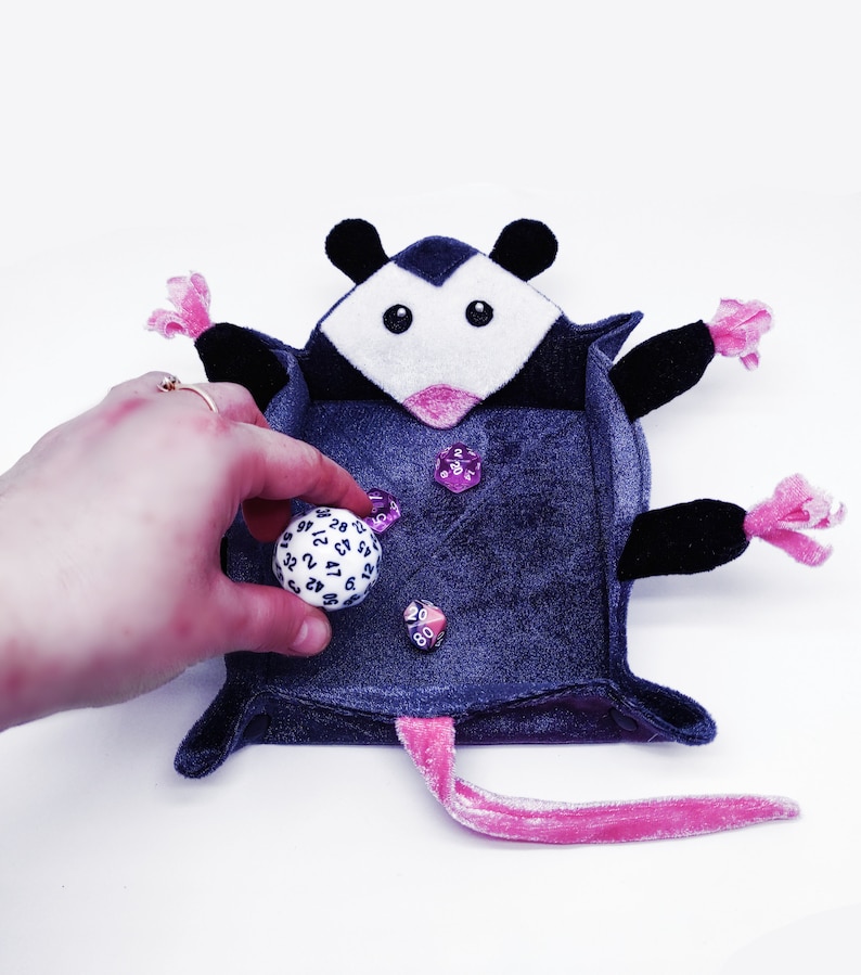 May include: A plush grey and pink opossum dice tray with a pink nose and black eyes. The tray is filled with dice, including a white 20-sided die and several purple dice.