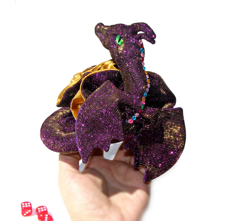 May include: A purple and gold glittery dragon plush toy with a colorful beaded necklace. The dragon has green eyes and is sitting on a hand.
