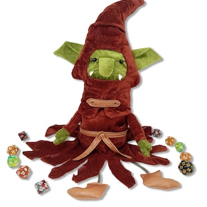 May include: A plush goblin figure in a burgundy hooded robe with green ears and face. The figure has tan leather-like feet and arms. Assorted dice are scattered around the base of the figure, suggesting a tabletop gaming theme.