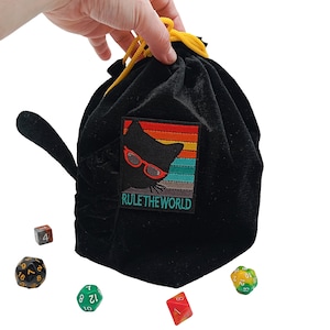 May include: Black velvet dice bag with a cat silhouette wearing sunglasses and the text "RULE THE WORLD" embroidered on the front. A yellow drawstring secures the top. Several multi-sided dice are scattered around the bag.