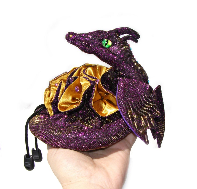 May include: A purple and gold glittery dragon-shaped pouch with a green eye. The pouch is closed and has a drawstring closure.