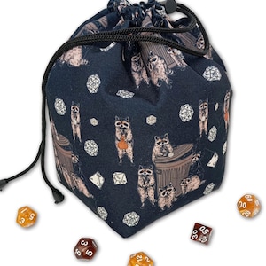 May include: A navy blue drawstring dice bag featuring a repeating pattern of raccoons and dice. The bag has a black drawstring closure and is surrounded by several multi-sided dice in various colors. The raccoons are depicted in a cartoon style.