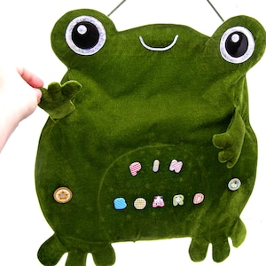 May include: A green plush frog-shaped pin board with a smiling face and large eyes. The board has a variety of colorful buttons and the word "PIN BOARD" is spelled out in colorful letters.