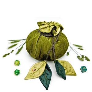 Puede incluir: Bolsa de dados de terciopelo verde con cierre de cord&oacute;n y dos borlas en forma de hoja verde y dorada. La bolsa est&aacute; sobre una superficie blanca con dos dados verdes y un dado verde y dorado.