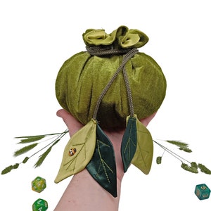 Puede incluir: Una bolsa de dados de terciopelo verde con cierre de cord&oacute;n y dos acentos de hojas verdes. La bolsa se sostiene en una mano con dos dados verdes delante.