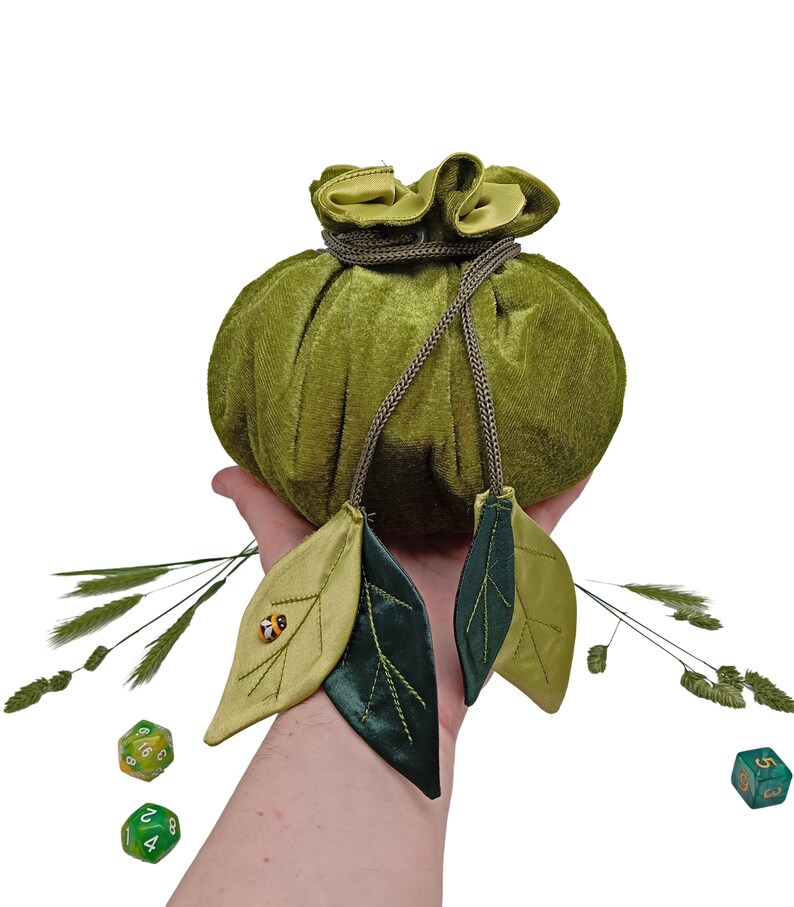 Puede incluir: Una bolsa de dados de terciopelo verde con cierre de cord&oacute;n y dos acentos de hojas verdes. La bolsa se sostiene en una mano con dos dados verdes delante.