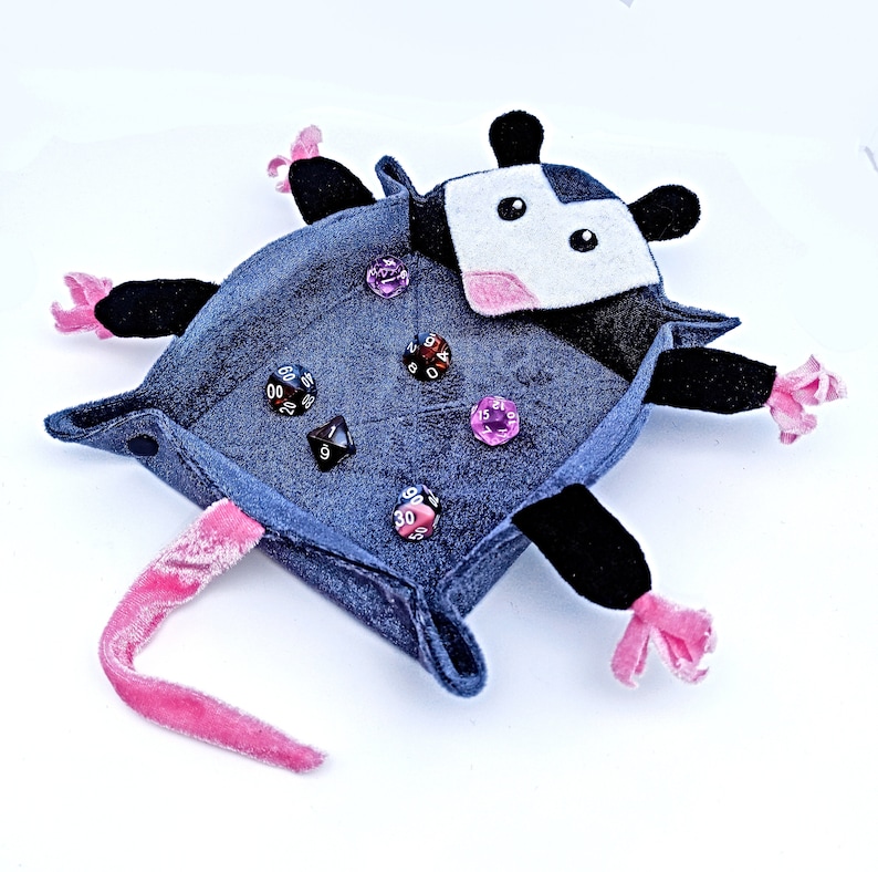 May include: A grey felt dice tray shaped like a possum with pink ears and a white face. The tray has a pink tail and black feet. There are several dice inside the tray, including a black d20, a black d6, a black d4, a pink d6, and a pink d12.