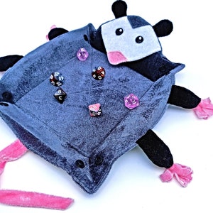 May include: A grey plush dice tray shaped like a possum with pink accents and black eyes. The tray is filled with various coloured dice.