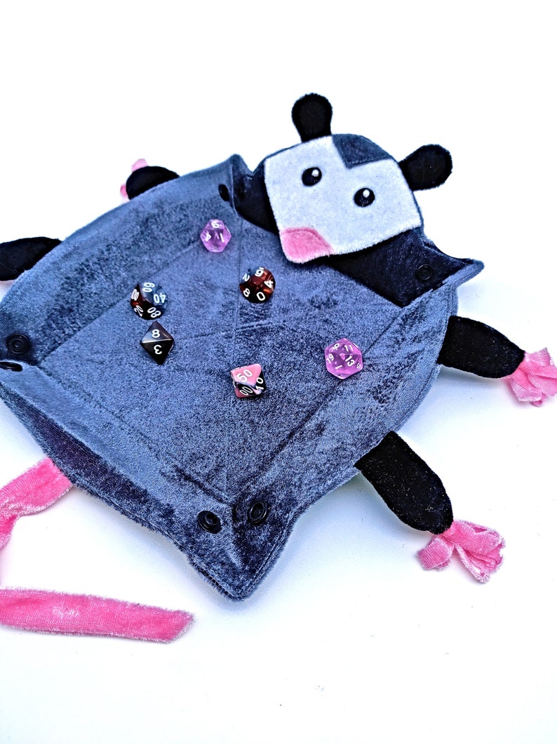 May include: A grey plush dice tray shaped like a possum with pink accents and black eyes. The tray is filled with various coloured dice.