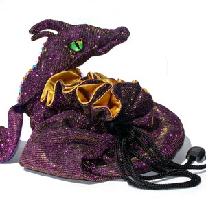 May include: Purple and gold glitter drawstring bag with a dragon head design. The bag has a black drawstring and a green eye.