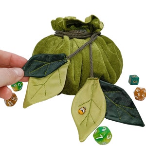 Puede incluir: Bolsa de dados de terciopelo verde con cierre de cord&oacute;n y solapa en forma de hoja. La bolsa est&aacute; llena de dados de colores y tiene una peque&ntilde;a mariquita en una de las solapas.
