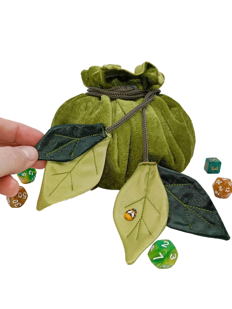 Puede incluir: Bolsa de dados de terciopelo verde con cierre de cord&oacute;n y solapa en forma de hoja. La bolsa est&aacute; llena de dados de colores y tiene una peque&ntilde;a mariquita en una de las solapas.